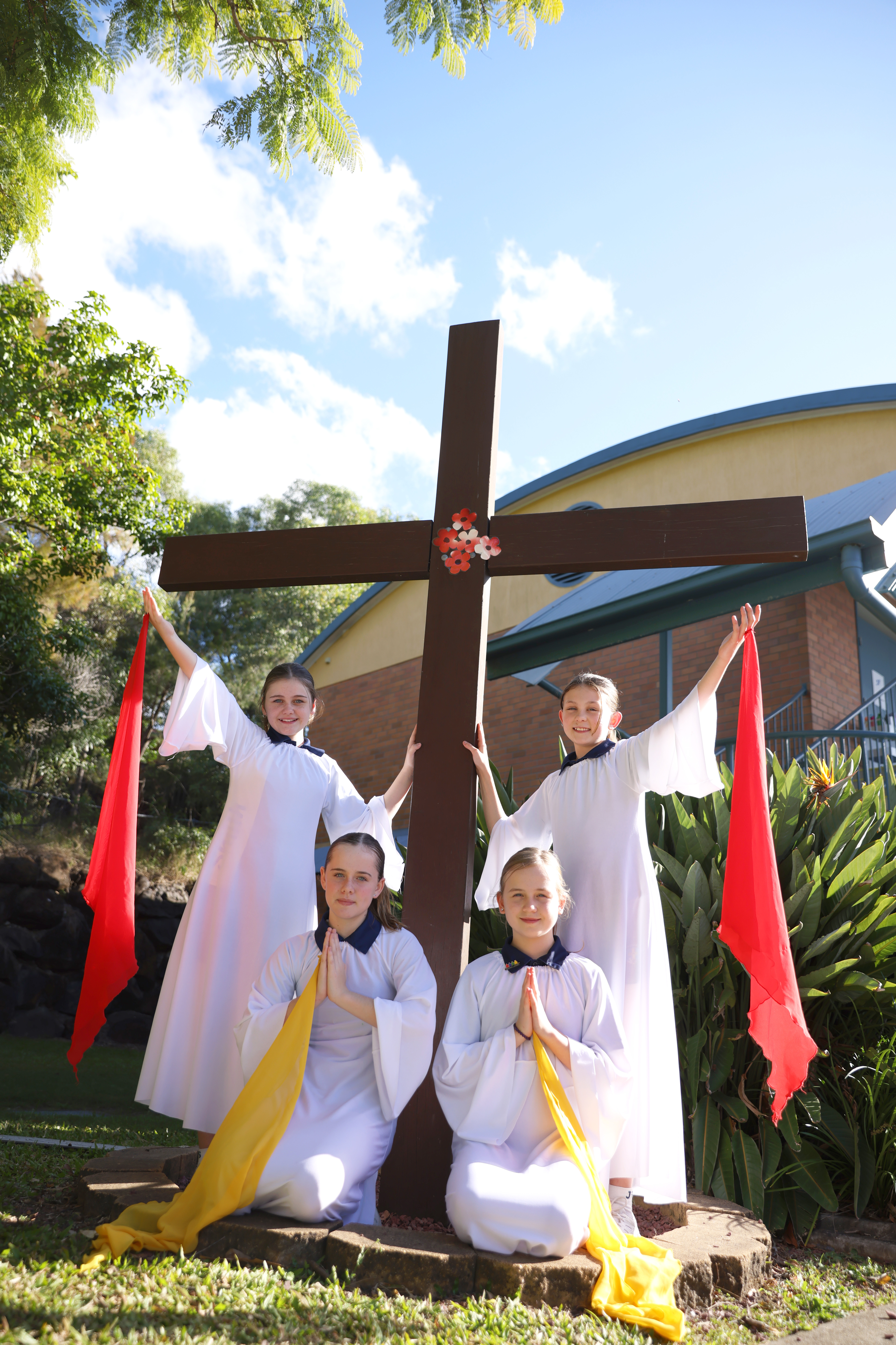 Students dressed in white robes gather around a large wooden cross on the school grounds, holding red and yellow fabric banners