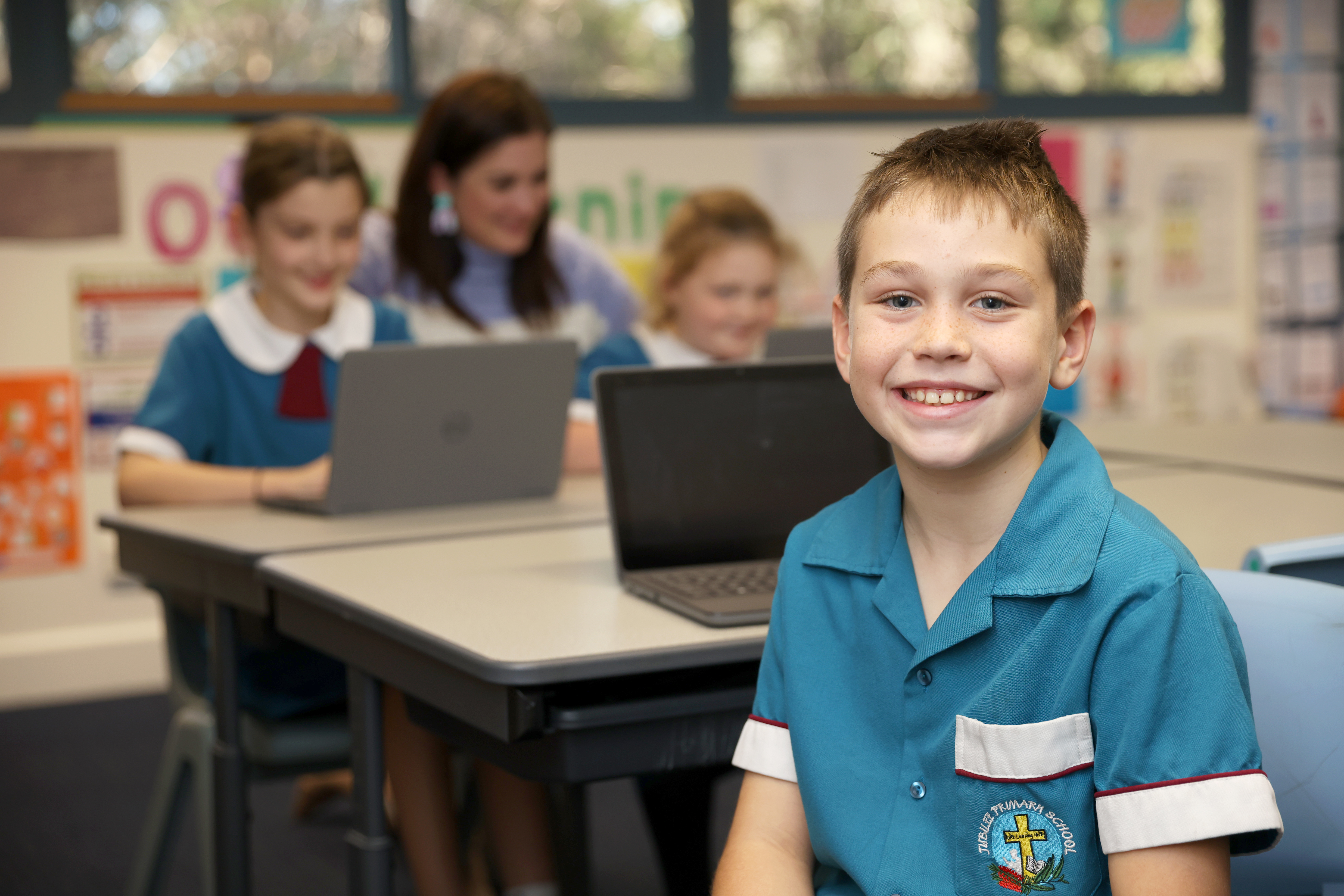 Students seated at desks in a classroom using laptops for learning, wearing school uniforms, with classroom displays visible in