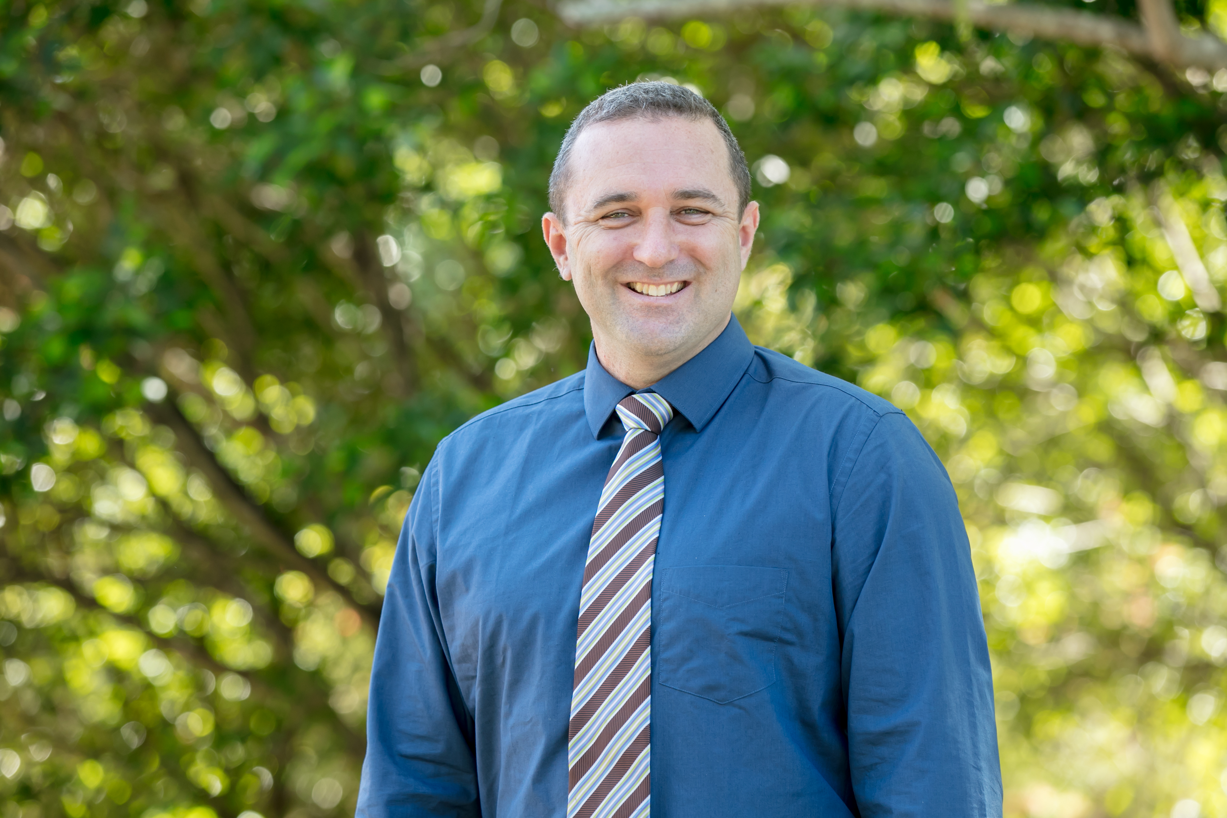 Staff member standing outdoors on school grounds, wearing a blue shirt and striped tie, with trees and greenery in the backgroun