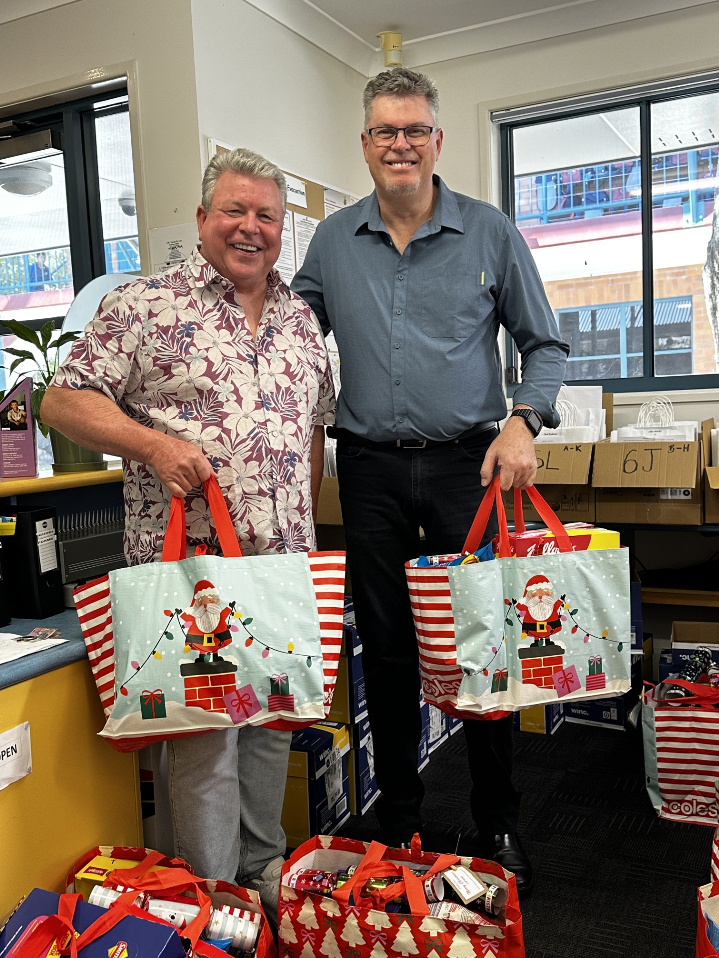 Staff members standing indoors holding festive gift bags, surrounded by donated items prepared for a school community initiative